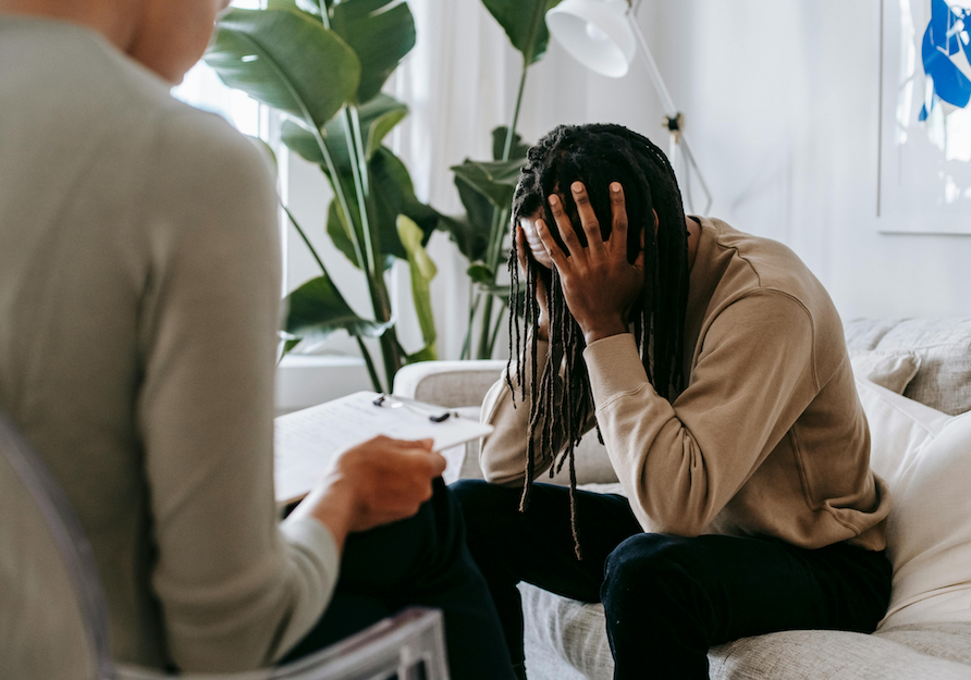 Eine Person sitzt auf einem Sofa und hält sich die Hände vors Gesicht. Ihr gegenüber sitzt eine weitere Person, die ein Klemmbrett hält – Szene aus einem Beratungs- oder Therapiegespräch.
