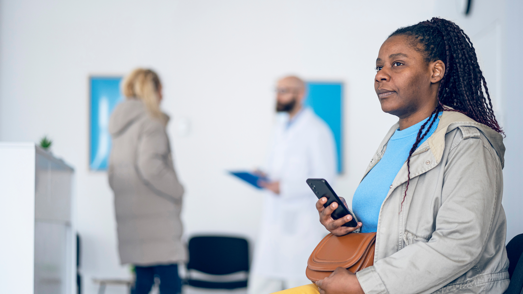 Eine Frau mittleren Alters sitzt mit dem Smartphone in der Hand im Wartezimmer einer Arztpraxis, im Hintergrund spricht ein Arzt mit einer Patientin.
